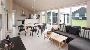 A spacious living area with a wooden floor, a dining table with chairs, a sofa with cushions, and large windows offering a view of the outdoors.