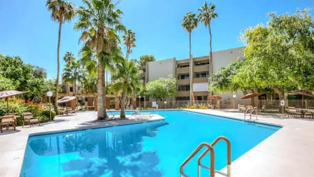 Ein Swimmingpool mit Palmen und Liegestühlen, umgeben von einem mehrstöckigen Gebäude mit Balkonen.