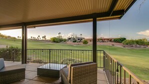Een overdekt terras met rieten meubilair en een glazen tafel, met uitzicht op een golfbaan en huizen op de achtergrond.