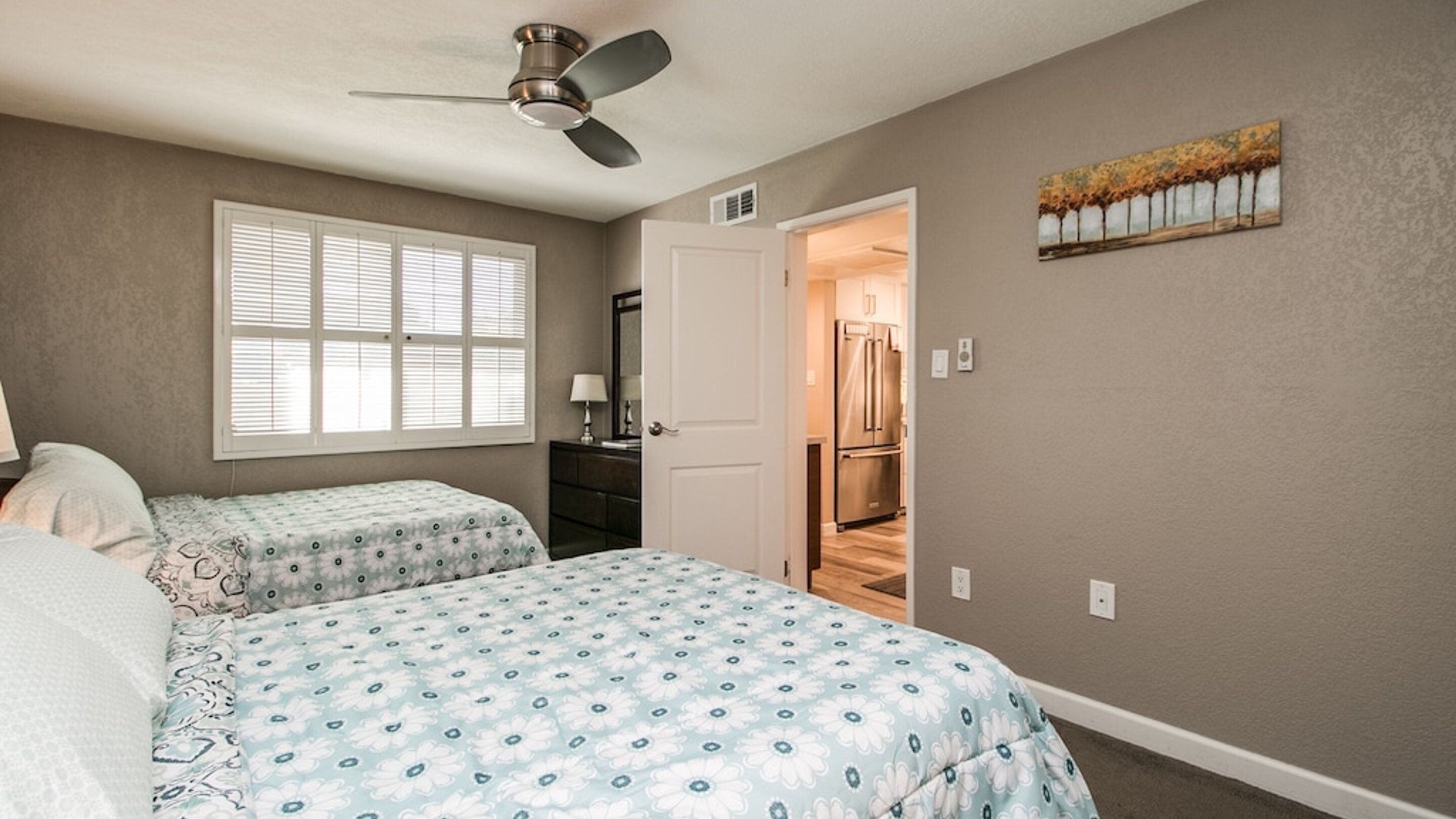 A bedroom with two beds, a ceiling fan, a window with blinds, a door leading to a hallway, and a painting on the wall.