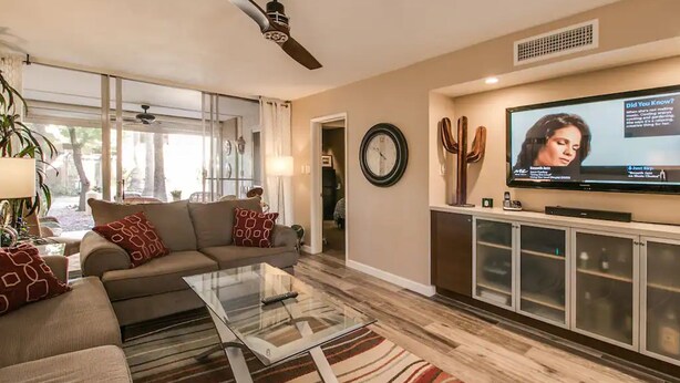 A modern living room with a glass coffee table, a sofa with patterned cushions, a television mounted on a cabinet, and a large clock on the wall. - Cozy 2-bdrm Condo in Heart of Old Town Scottsdale! (Scottsdale)