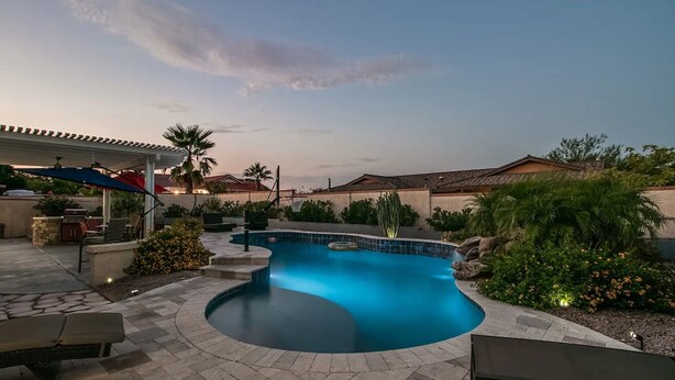 Ein Pool mit geschwungener Form, umgeben von einer Terrasse mit Gartenmöbeln und einem überdachten Sitzbereich.
