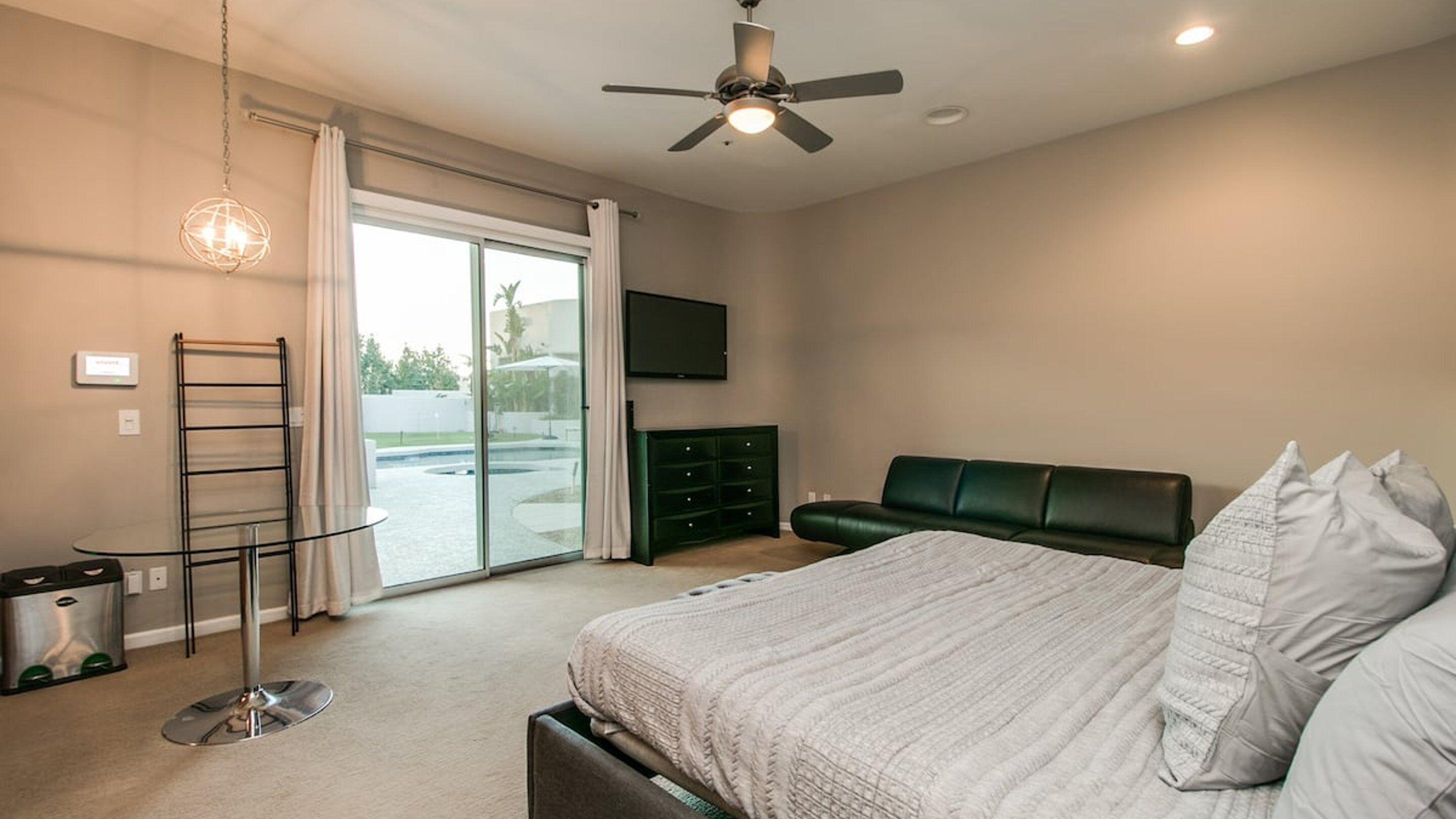 Une chambre avec un lit, un canapé vert, une télévision, un ventilateur de plafond et une porte-fenêtre coulissante donnant sur une zone avec piscine.