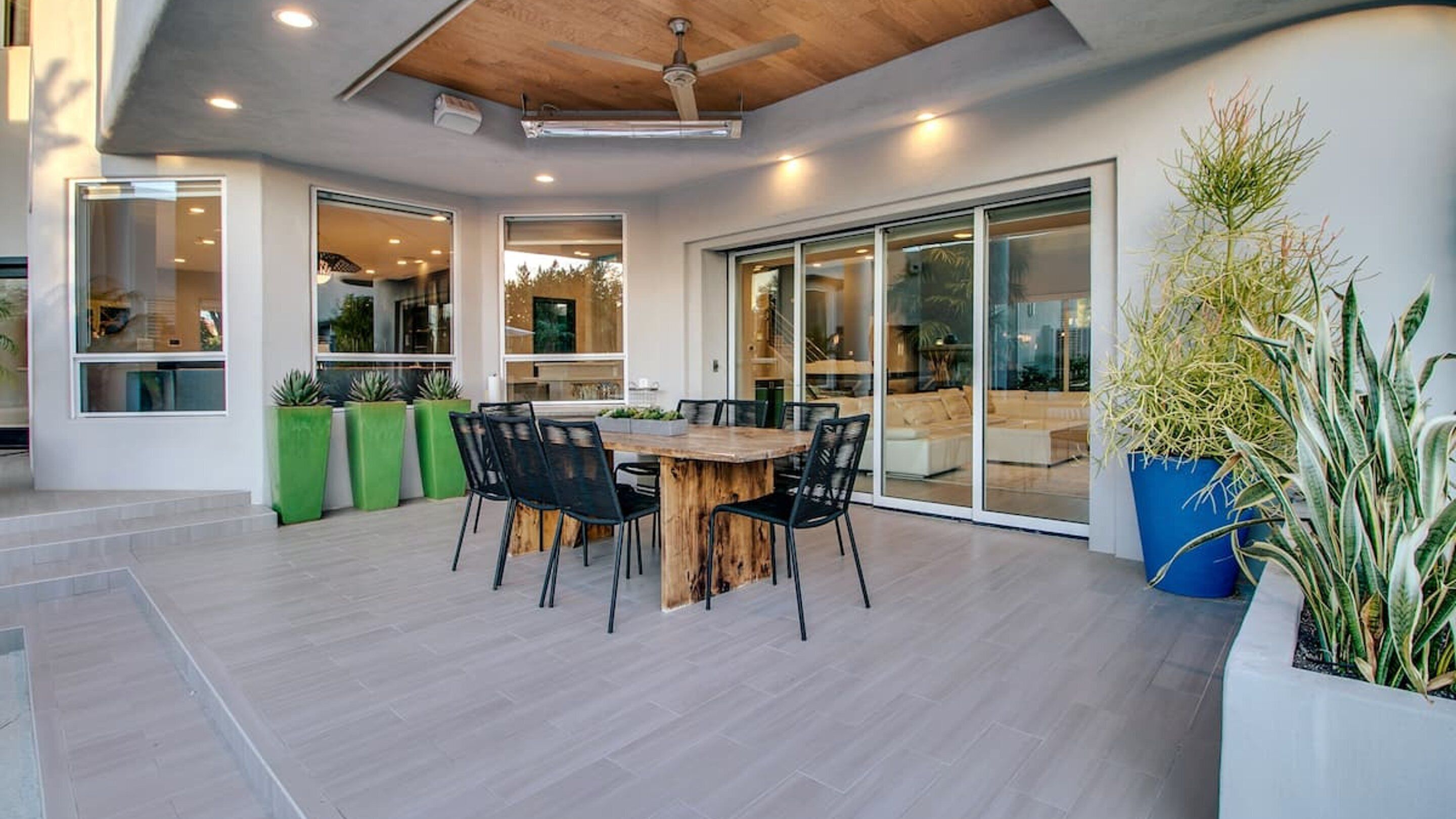 A modern outdoor dining area with a wooden table and chairs, surrounded by potted plants and a large blue planter.