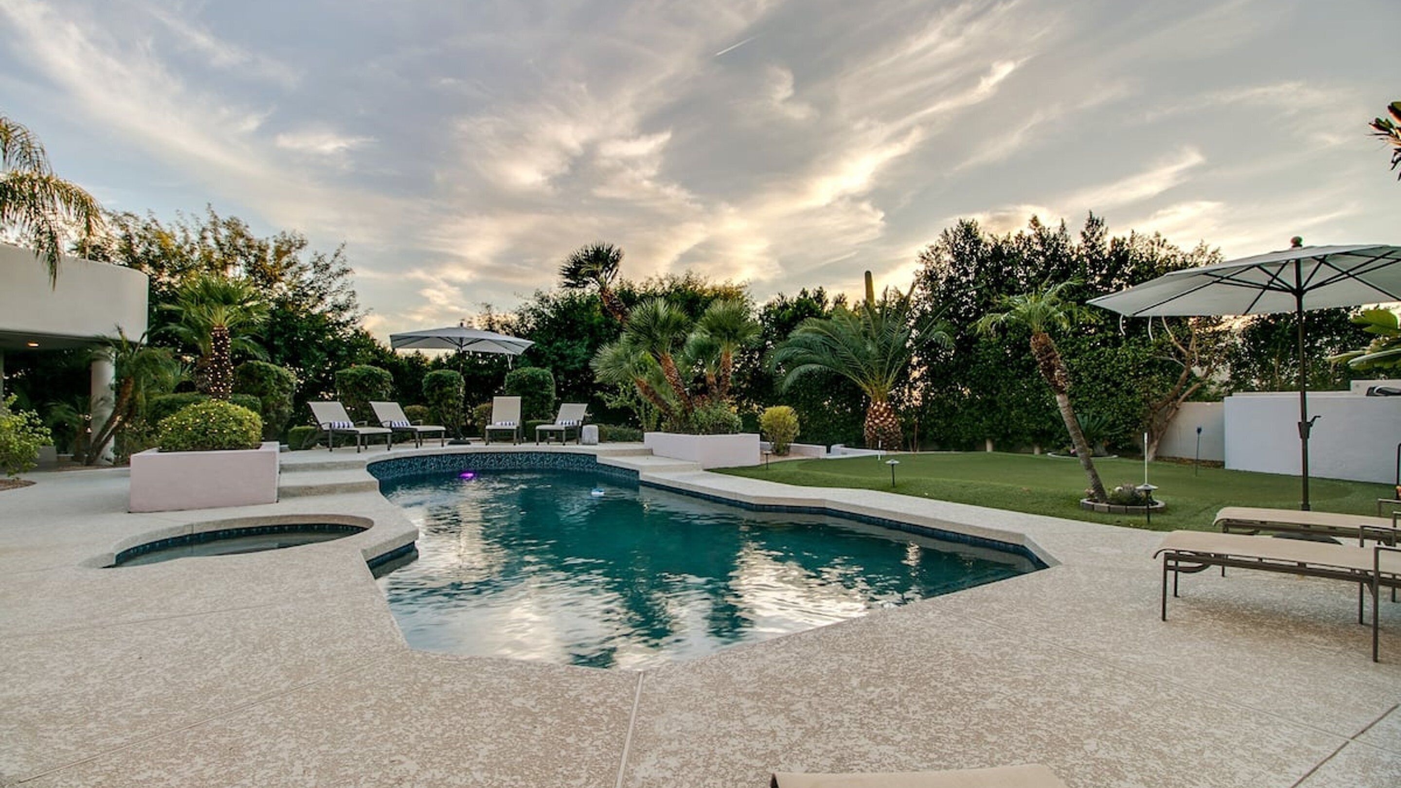 A swimming pool with a curved edge, surrounded by lounge chairs and umbrellas, set in a landscaped garden with various plants and trees.