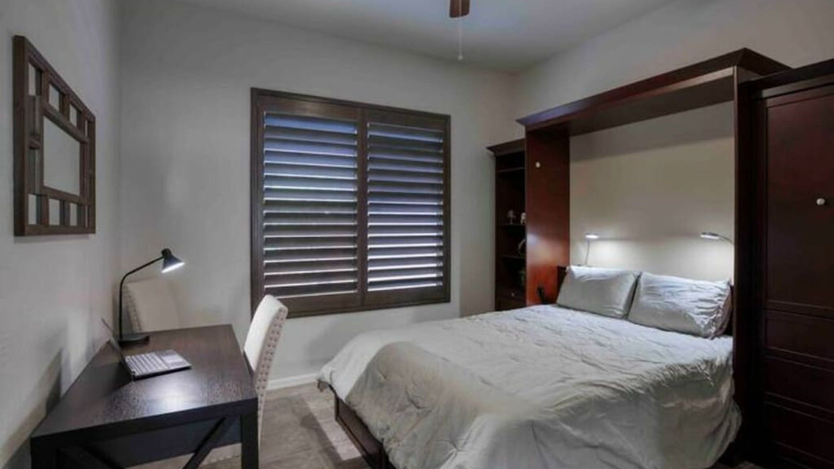 Un dormitorio con cama, escritorio, silla, ventana con persianas y ventilador de techo.