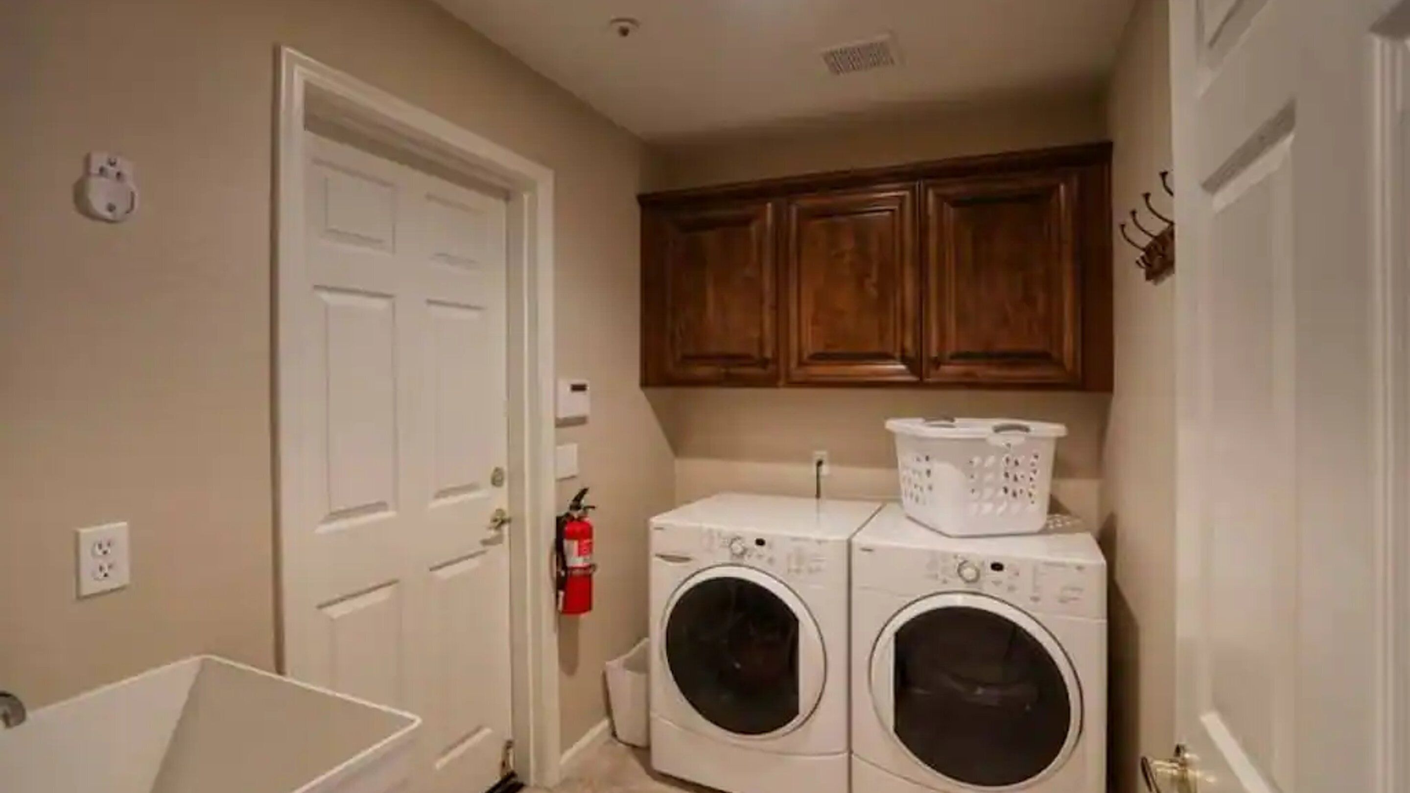 A laundry room with two front-loading washing machines, wooden cabinets above, a white laundry basket on top, and a white utility sink.