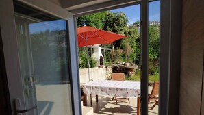 Une porte-fenêtre coulissante donne sur une terrasse extérieure équipée d’une table et de chaises, d’un parasol rouge et d’un mur en pierre.