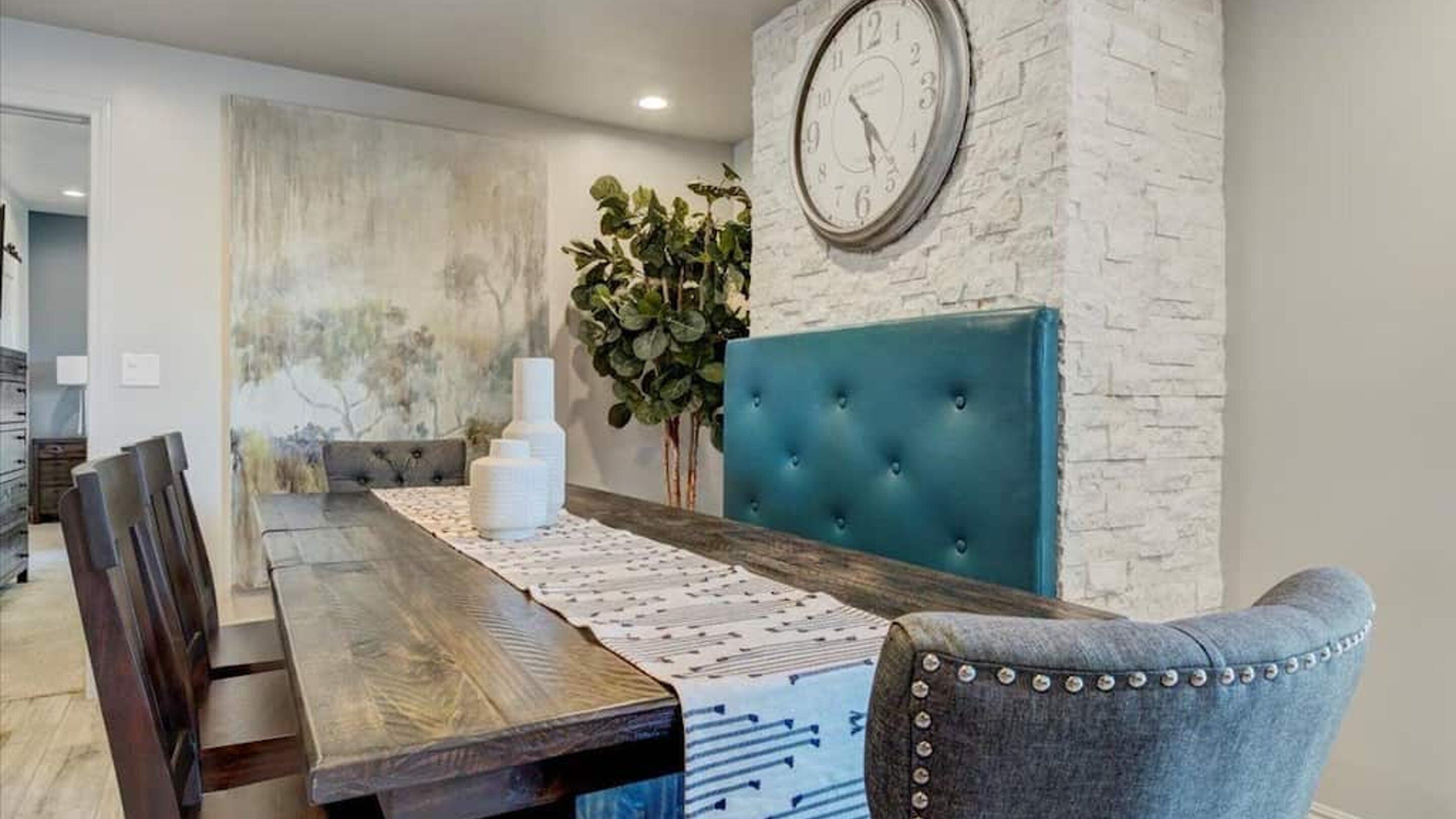 A dining area with a wooden table, a blue upholstered headboard, and a clock on the wall.