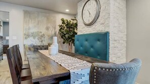 A dining area with a wooden table, a blue upholstered headboard, and a clock on the wall.