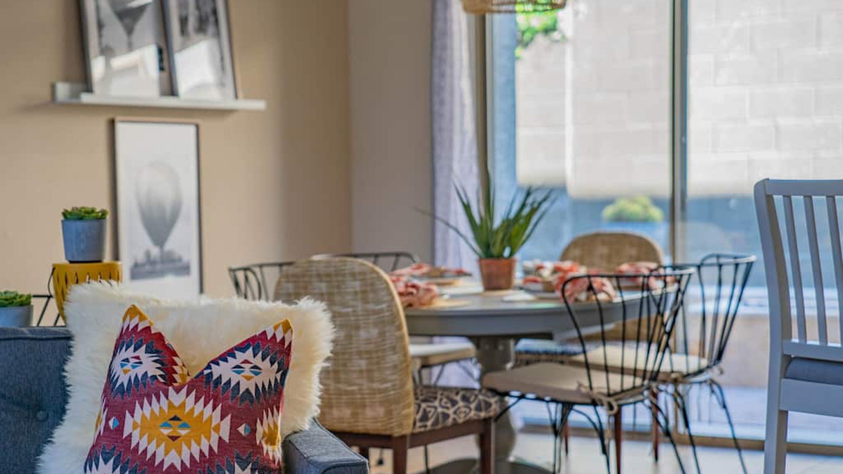 A dining area with a table set for two, surrounded by chairs, a potted plant, and a decorative pillow.