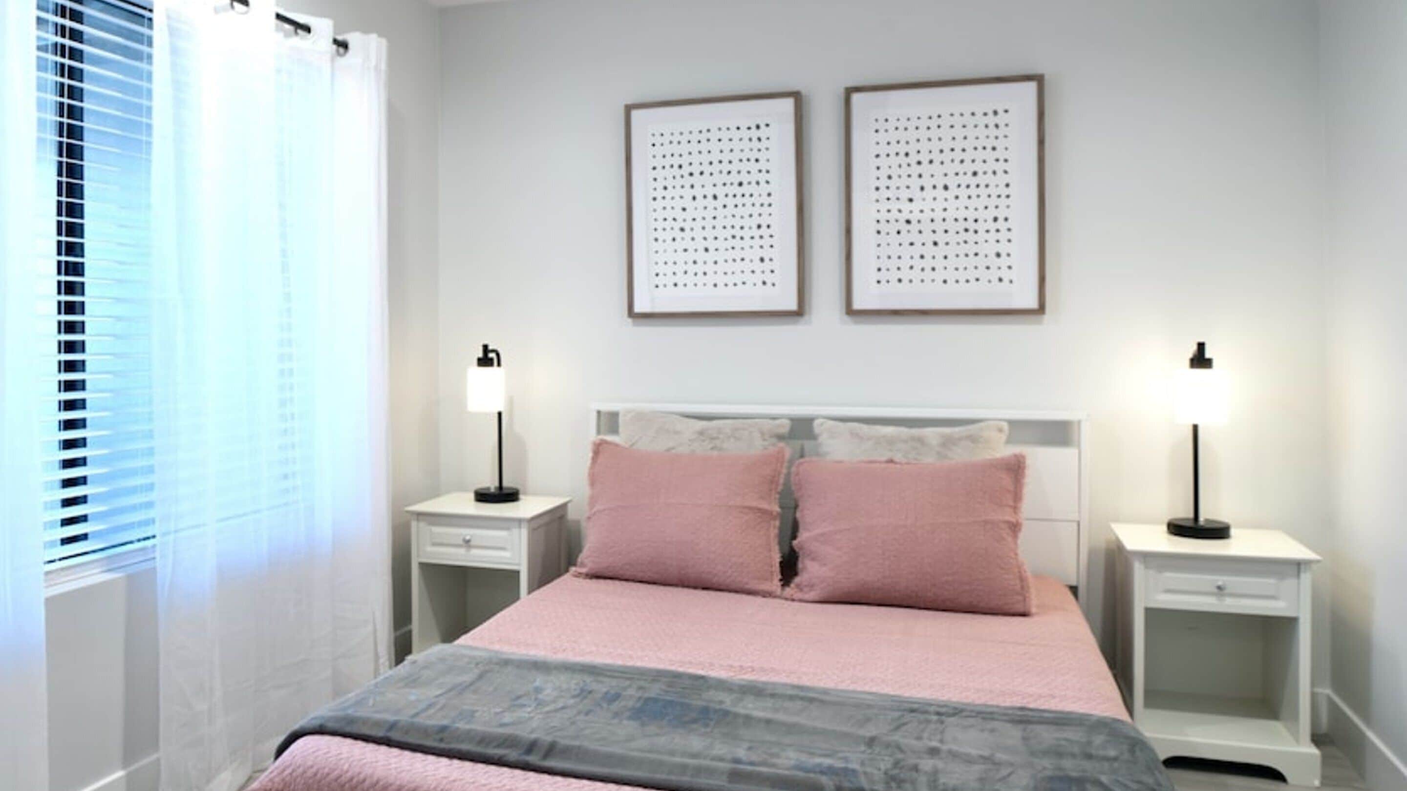 A bedroom with a bed, two bedside tables, a nightstand, a window with blinds, and two framed pictures on the wall.