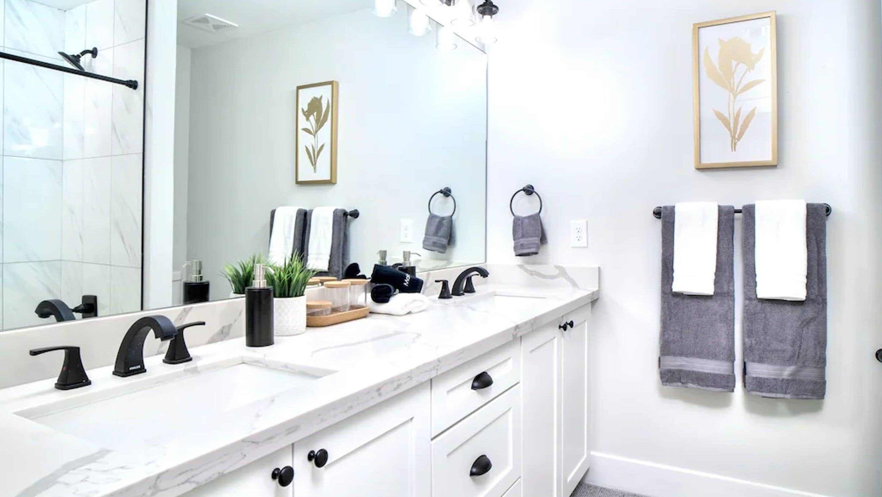 Un baño moderno con un lavamanos de mármol blanco, grifos negros y un espejo grande. Hay toallas colgadas en un perchero y un cuadro en la pared.