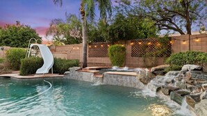 A swimming pool with a waterfall feature, a slide, and a brick wall with string lights. - Massive North Scottsdale 6 Bdrm w/ Oasis Backyard! (Scottsdale)