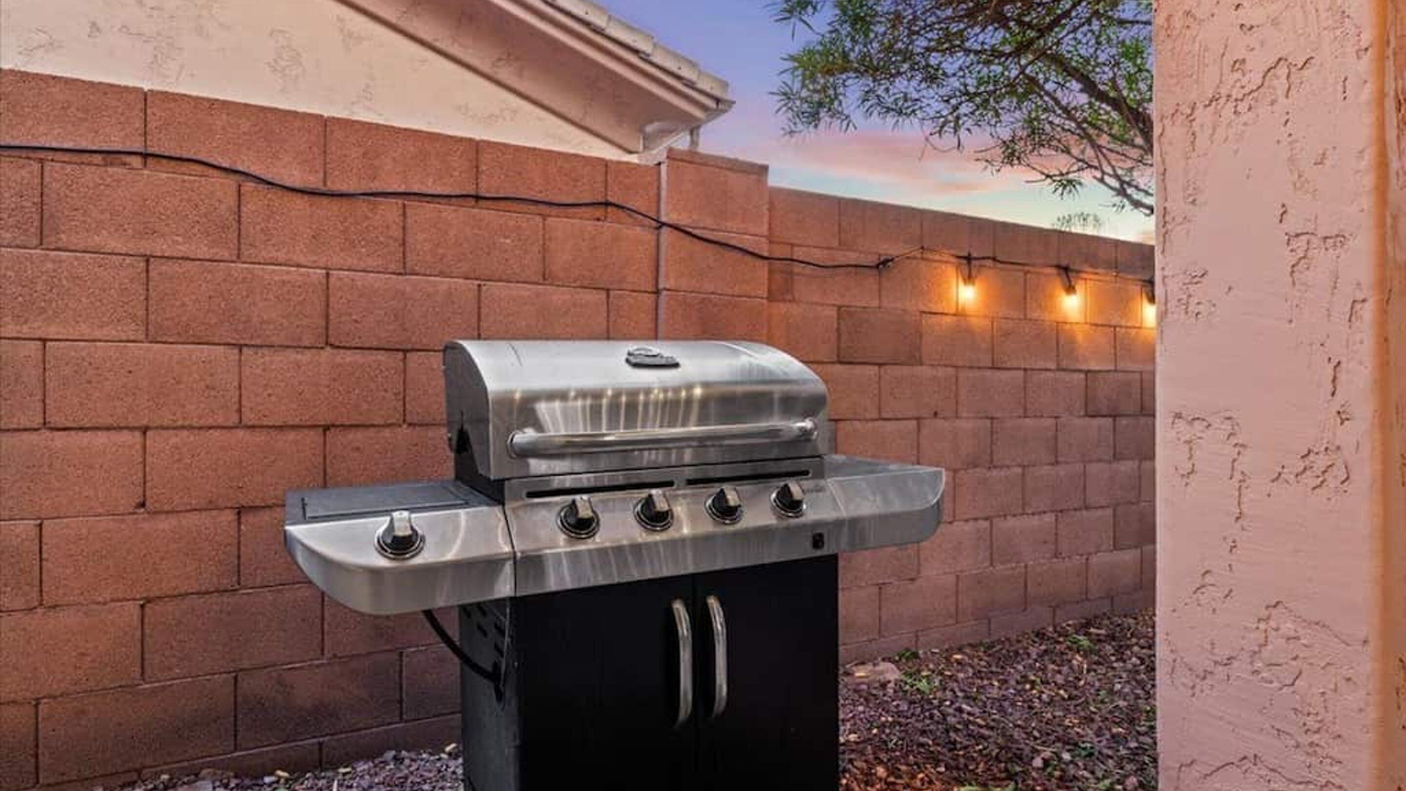 Un barbecue en acier inoxydable doté de quatre brûleurs et d’une étagère latérale, installé contre un mur de briques et agrémenté d’une guirlande lumineuse.