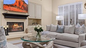 A living room with a fireplace, a sofa, a glass coffee table, and a painting of a desert landscape. - Massive North Scottsdale 6 Bdrm w/ Oasis Backyard! (Scottsdale)