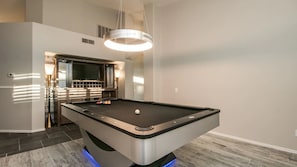Una mesa de billar moderna con luz LED integrada, un área de estar cómoda con sofá y televisor, y una lámpara colgante elegante sobre la mesa.