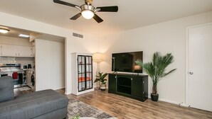 Un salon moderne avec un canapé gris, un téléviseur à écran plat, un ventilateur de plafond et une cuisine visible au fond.