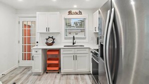 A modern kitchen with white cabinets, a stainless steel refrigerator, and a window with blinds.