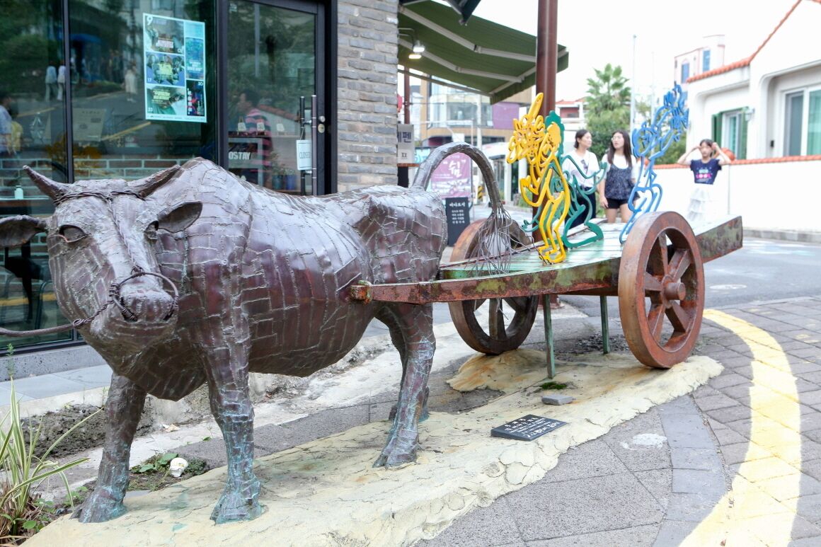 A metal sculpture of a bull with a cart, located on a sidewalk near a building with a glass storefront.