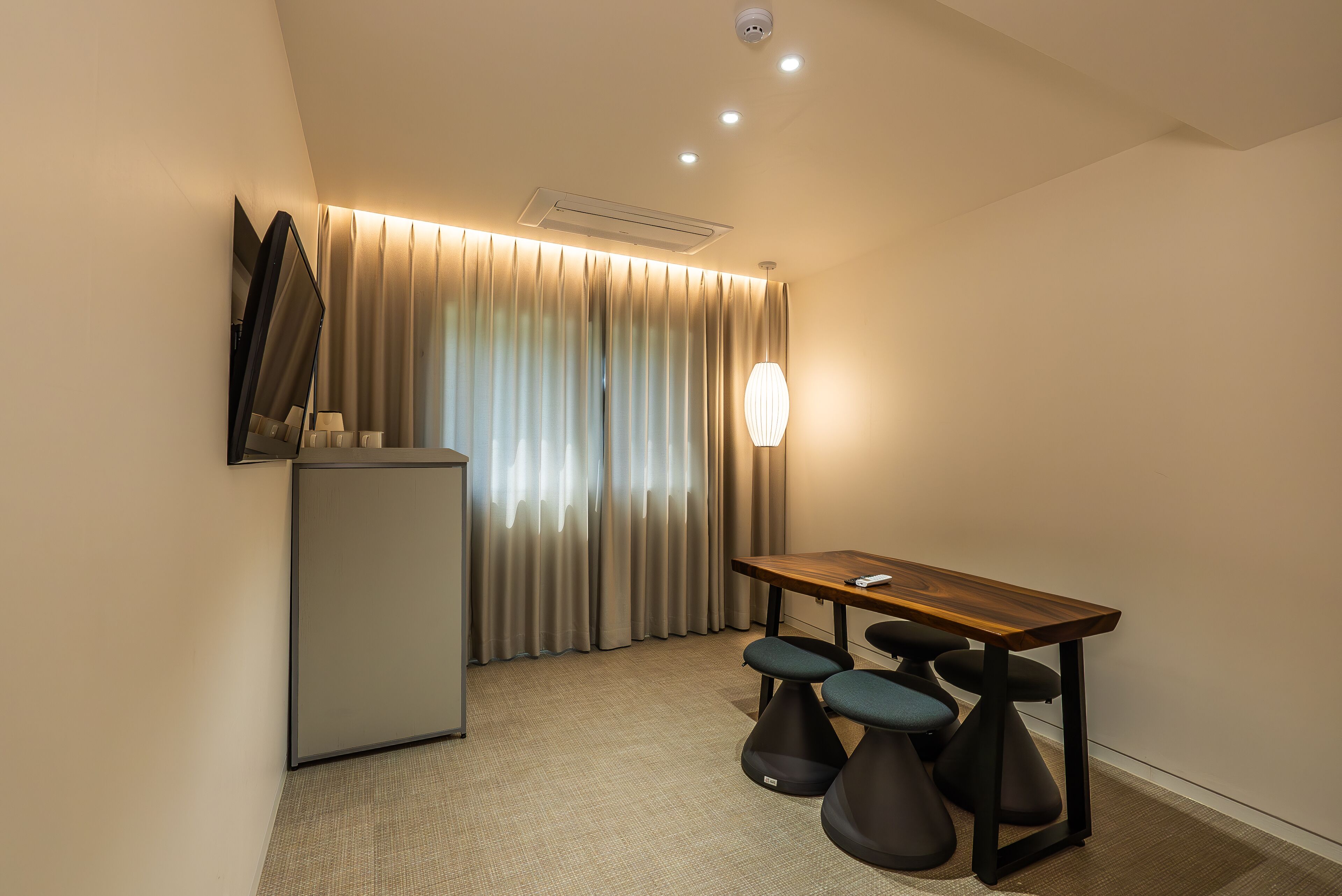 A hotel room with a desk, two stools, a flat-screen TV, a mini-fridge, and a large window with curtains.