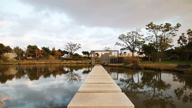 A long, straight concrete pathway leading to a structure with a grid-like facade, surrounded by trees and a body of water. - Hotel The Grang Seogwipo (Seogwipo)