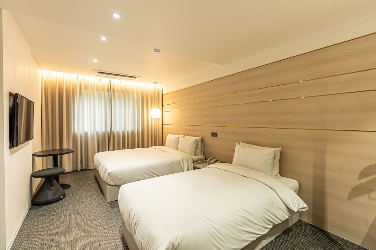 Een hotelkamer met twee bedden, een tafeltje, een stoel, een televisie en een gordijn.