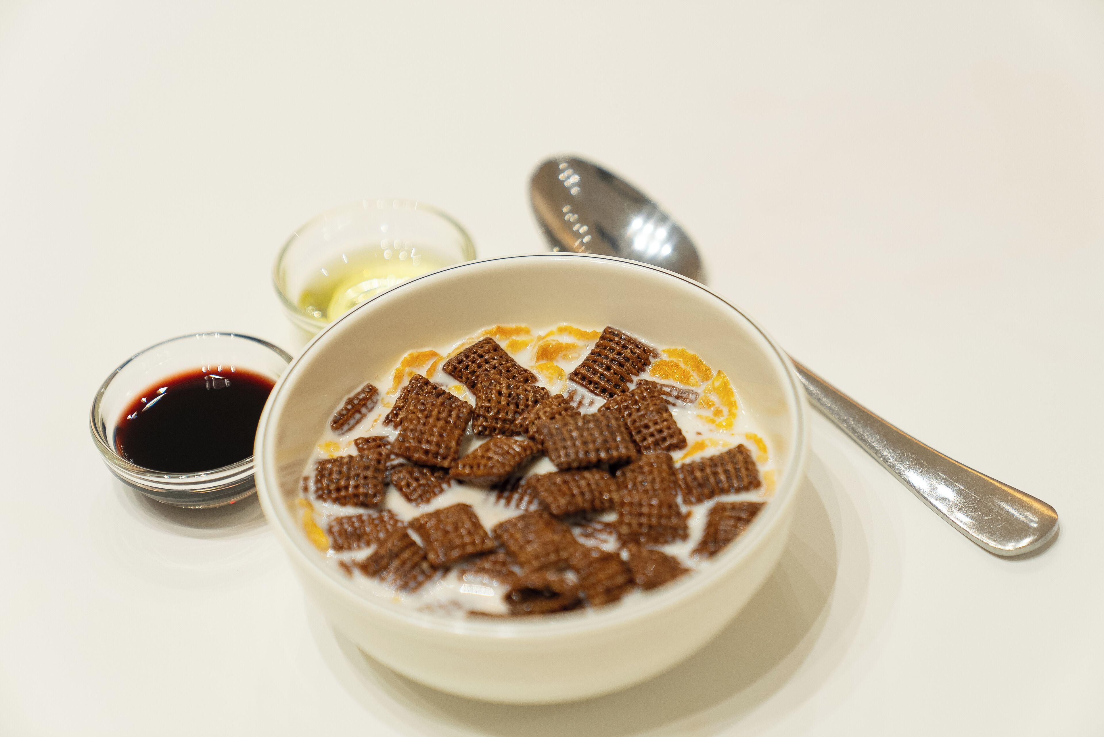 Un tazón de cereal con leche y pedazos de chocolate, un tazón pequeño de almíbar y una cuchara sobre una superficie blanca.