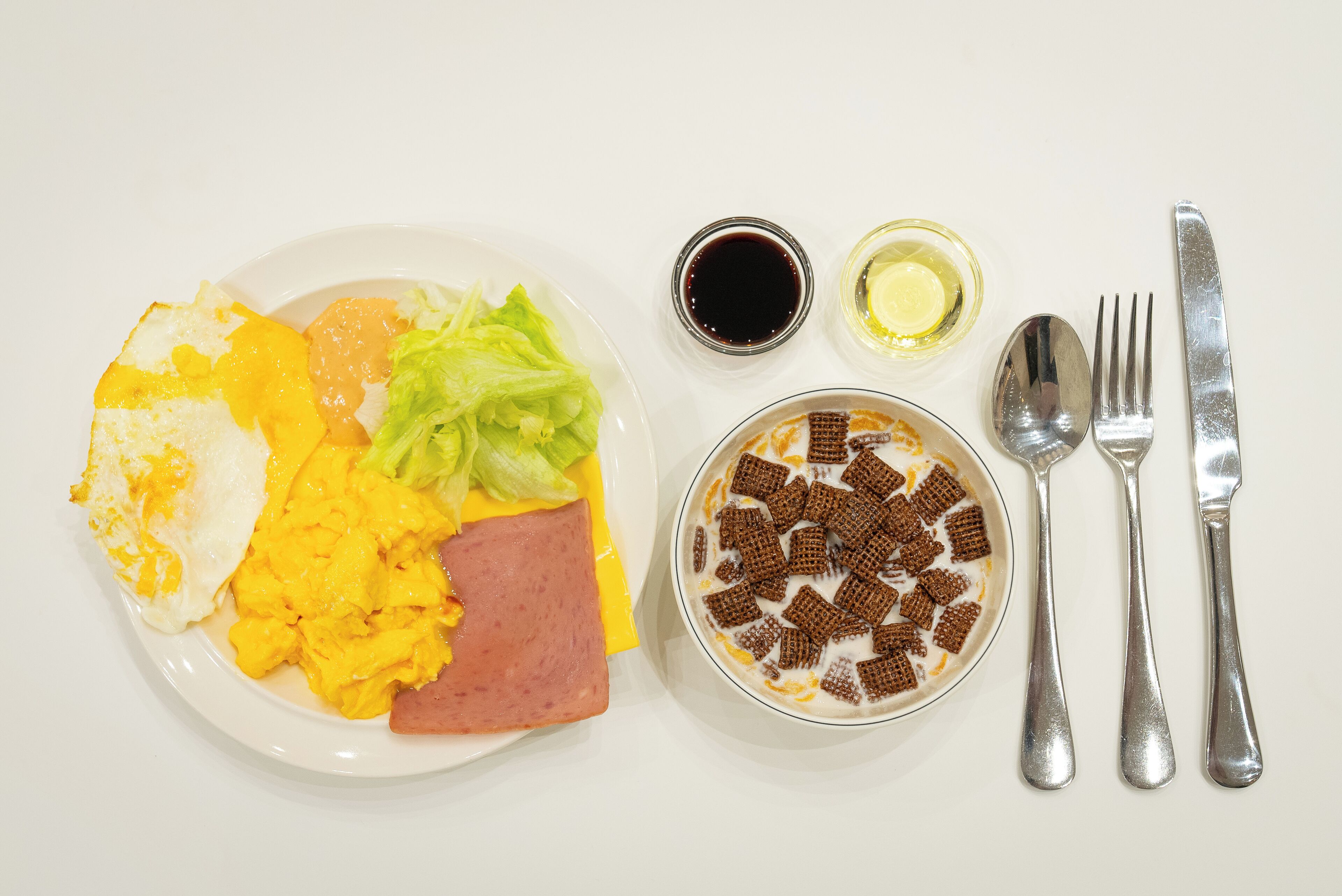 Ein Teller mit Rührei, Schinken und Salat, eine Schüssel mit Müsli und Milch sowie Gewürzen auf einem weißen Tisch.