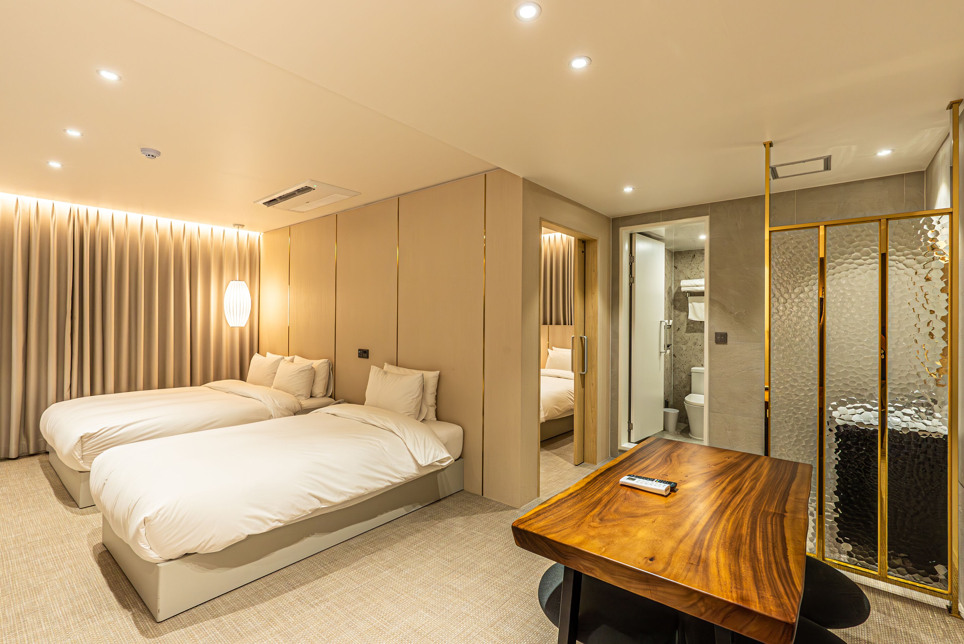 A hotel room with two beds, a wooden table, and a bathroom visible through a glass partition.