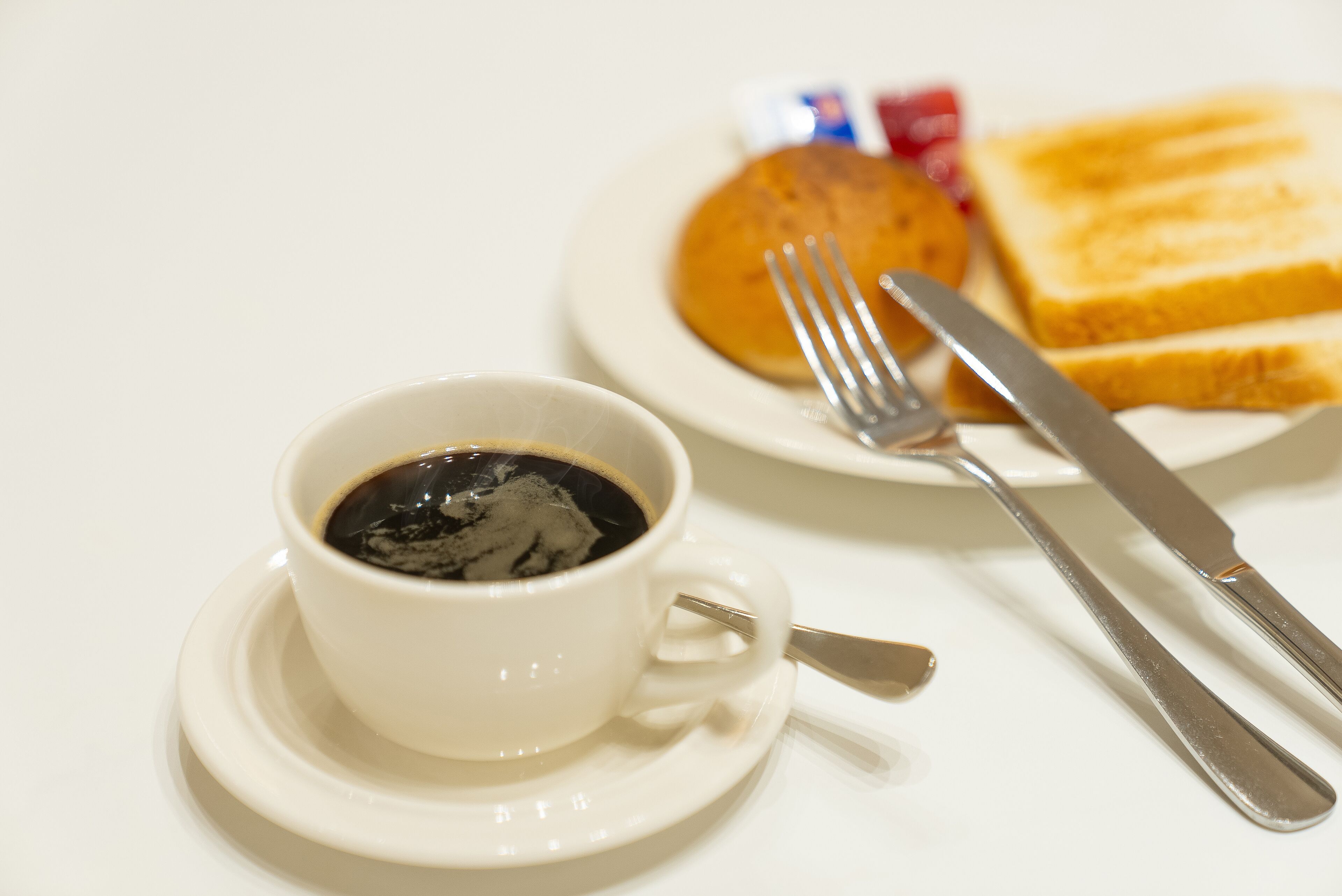 A white cup of coffee on a saucer with a fork and knife beside it.