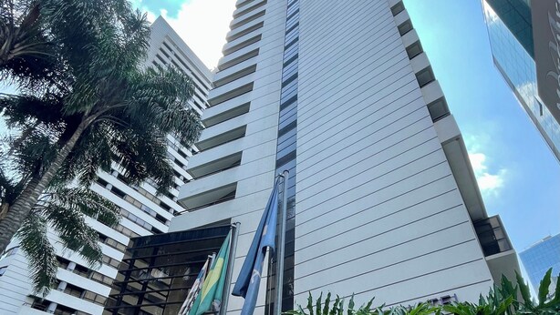 A modern building with a glass facade, surrounded by lush greenery and palm trees. - Três de Julho Flats - Paulista (São Paulo)