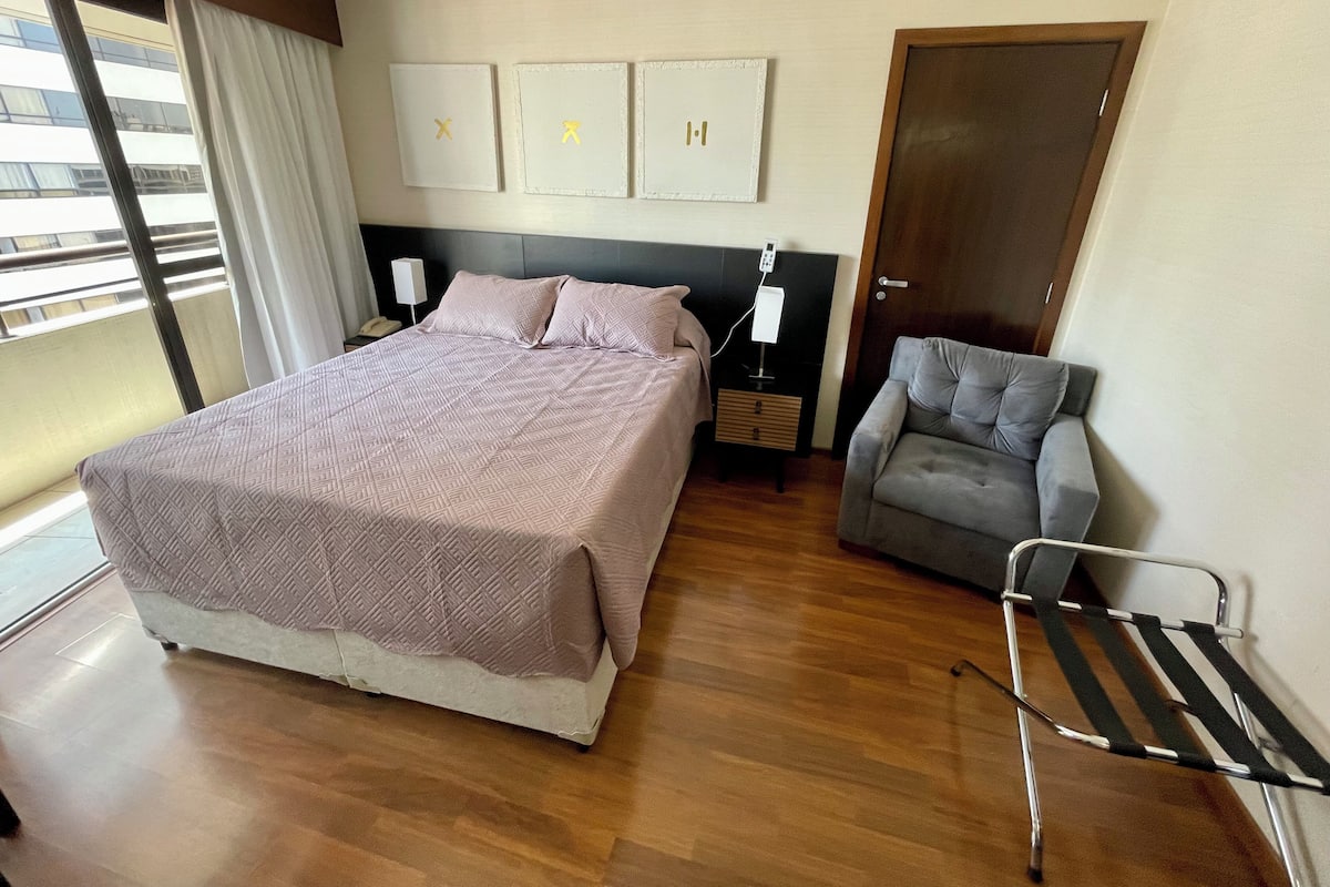 Une chambre à coucher moderne avec un grand lit, une table de chevet avec une lampe, un fauteuil gris et une fenêtre avec des rideaux.