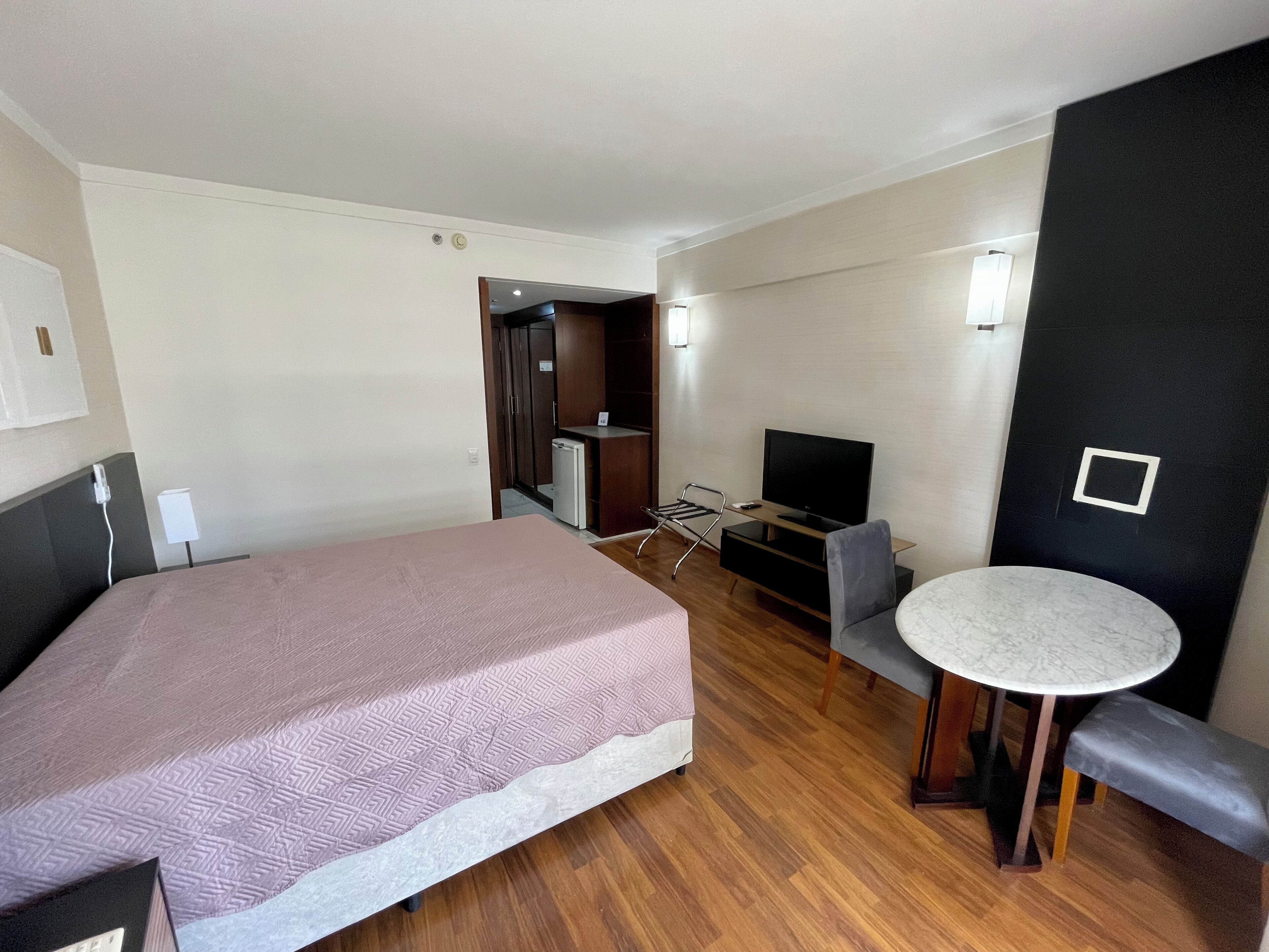 Et hotelværelse med en stor seng, et skrivebord med fjernsyn, et lille køleskab og et rundt bord med en bænk.