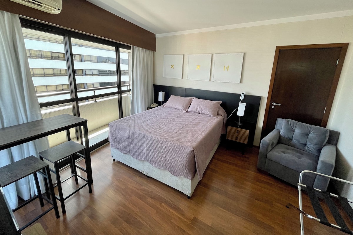 Une chambre d’hôtel moderne, dotée d’un grand lit, d’un bureau, d’une chaise et offrant une vue sur la ville grâce à de grandes fenêtres.