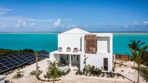 A modern white building with a balcony overlooking turquoise waters, solar panels, and tropical vegetation.