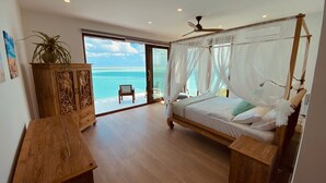 A bedroom with a bed, a wooden cabinet, a bench, and a view of the ocean.