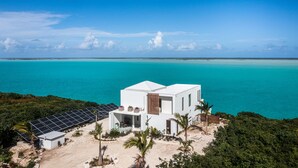 A modern white building with solar panels, surrounded by tropical vegetation and overlooking a turquoise sea.