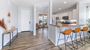A modern kitchen with white cabinets, a central island with bar stools, and a dining area with a table and chairs. - Boho Bungalow - 30ft pool & minutes from festivals  (Indio)