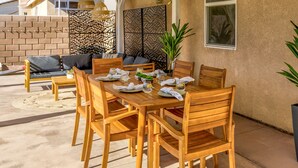 An outdoor dining area with wooden tables and chairs, a brick wall, and a decorative panel in the background. - Boho Bungalow - 30ft pool & minutes from festivals  (Indio)