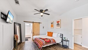 A bedroom with a bed, a ceiling fan, a TV mounted on the wall, and a small table with a clock. - Boho Bungalow - 30ft pool & minutes from festivals  (Indio)