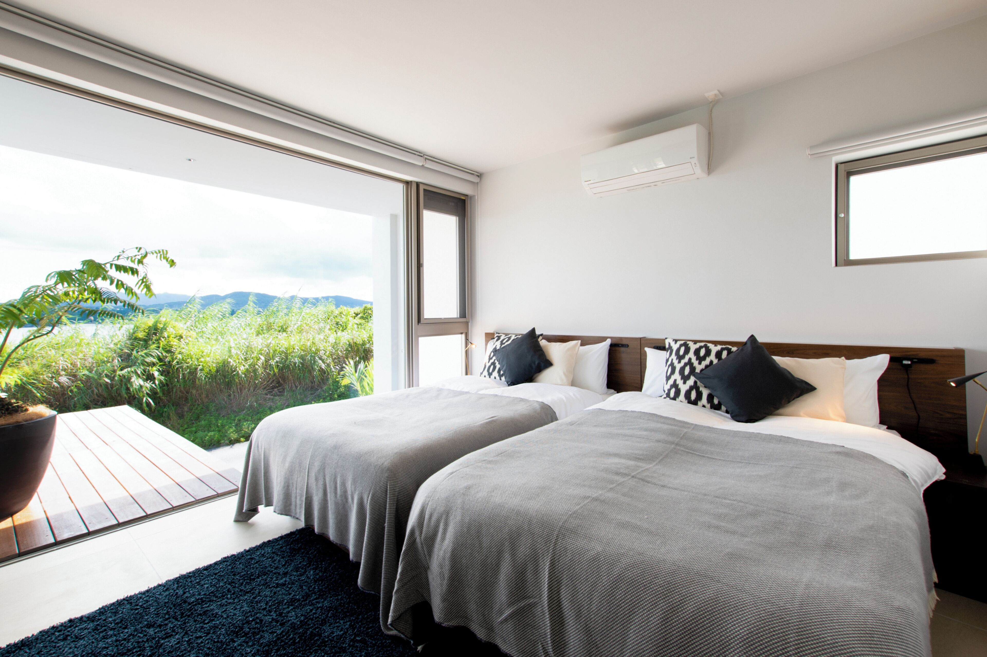 A modern hotel room with two beds, a large window, and a view of greenery.