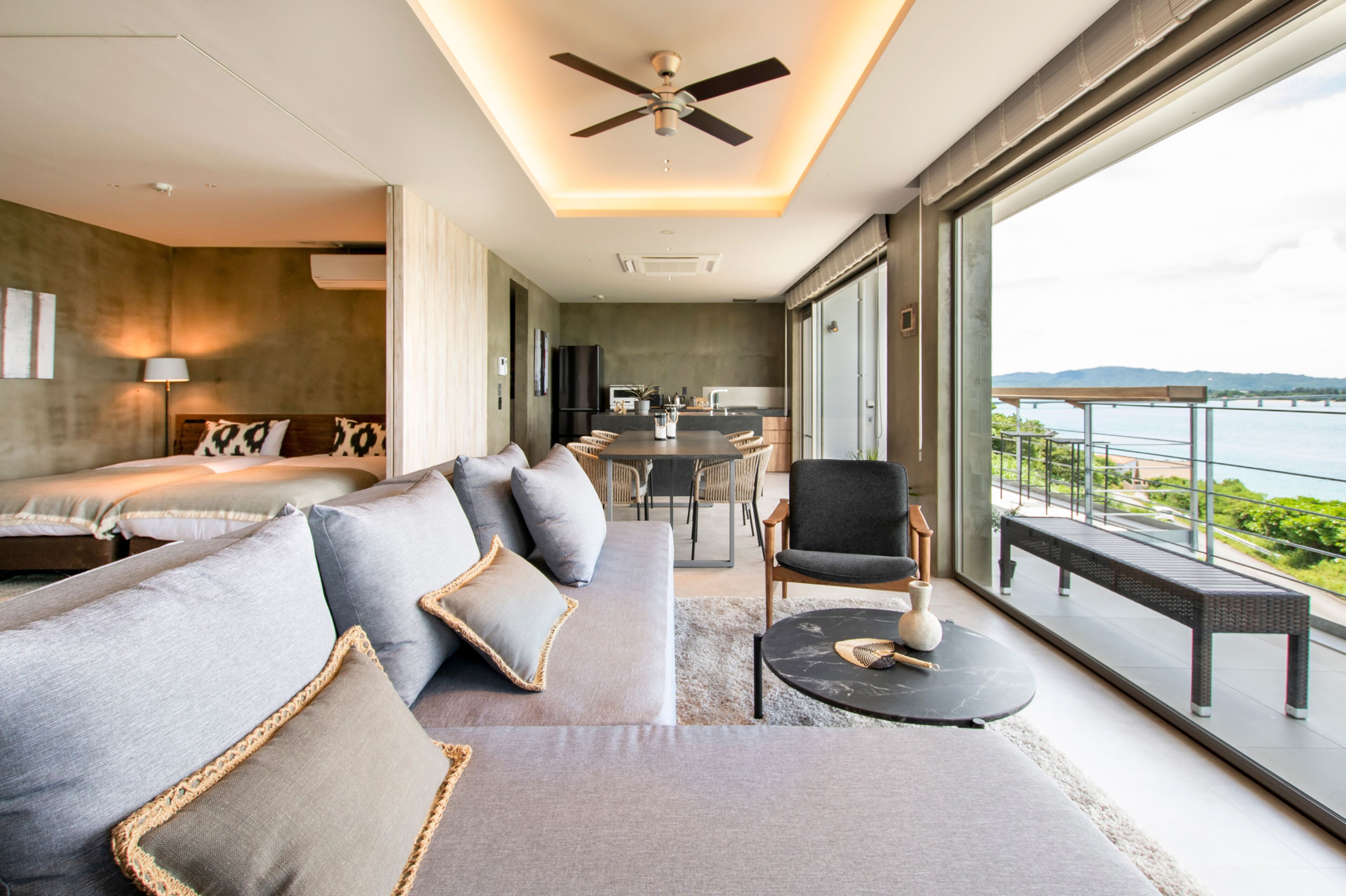 A modern hotel room with a large bed, a sofa, a dining area, and a balcony with a view of the sea.