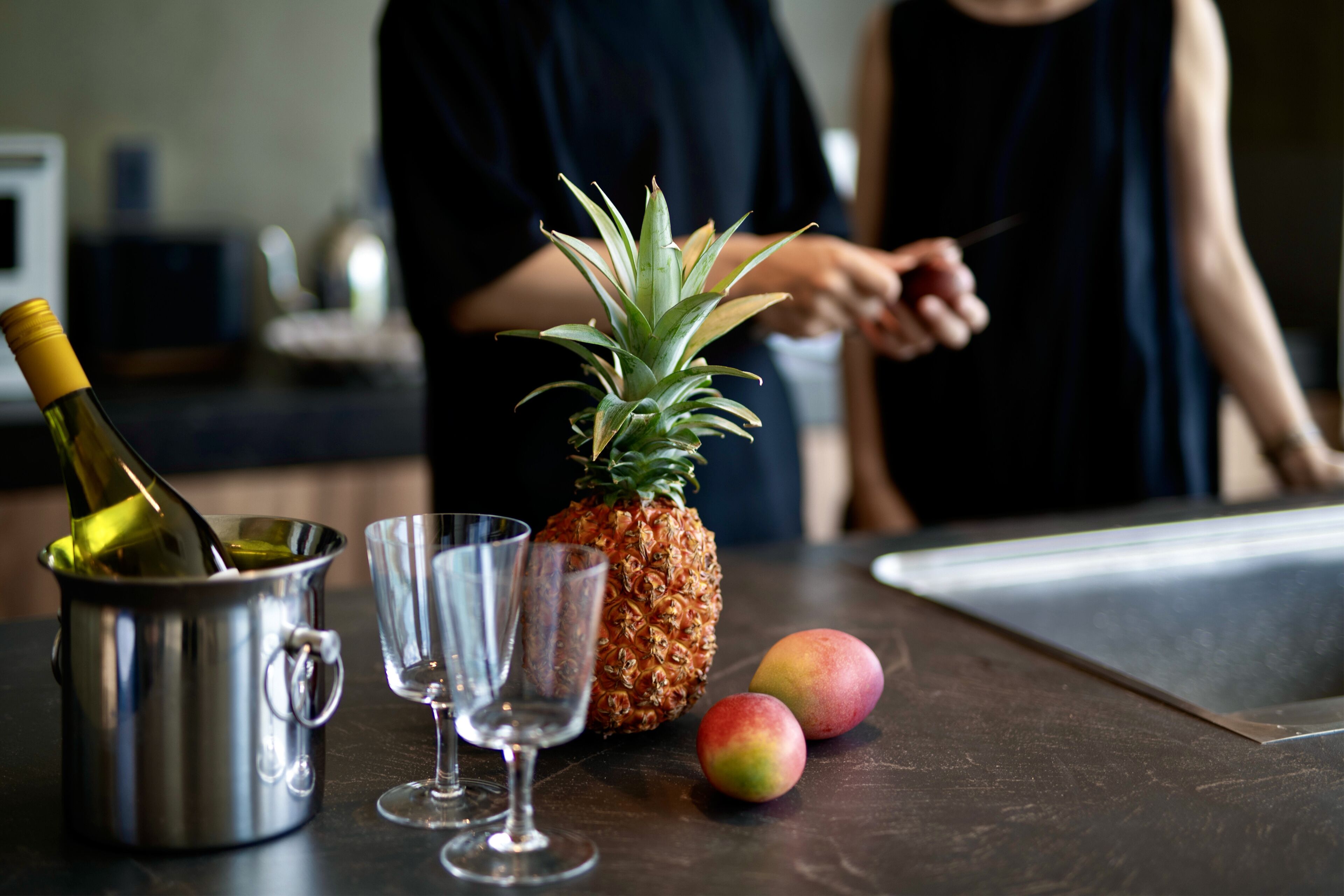 Una encimera de cocina con una botella de vino, dos copas, una piña y dos mangos.