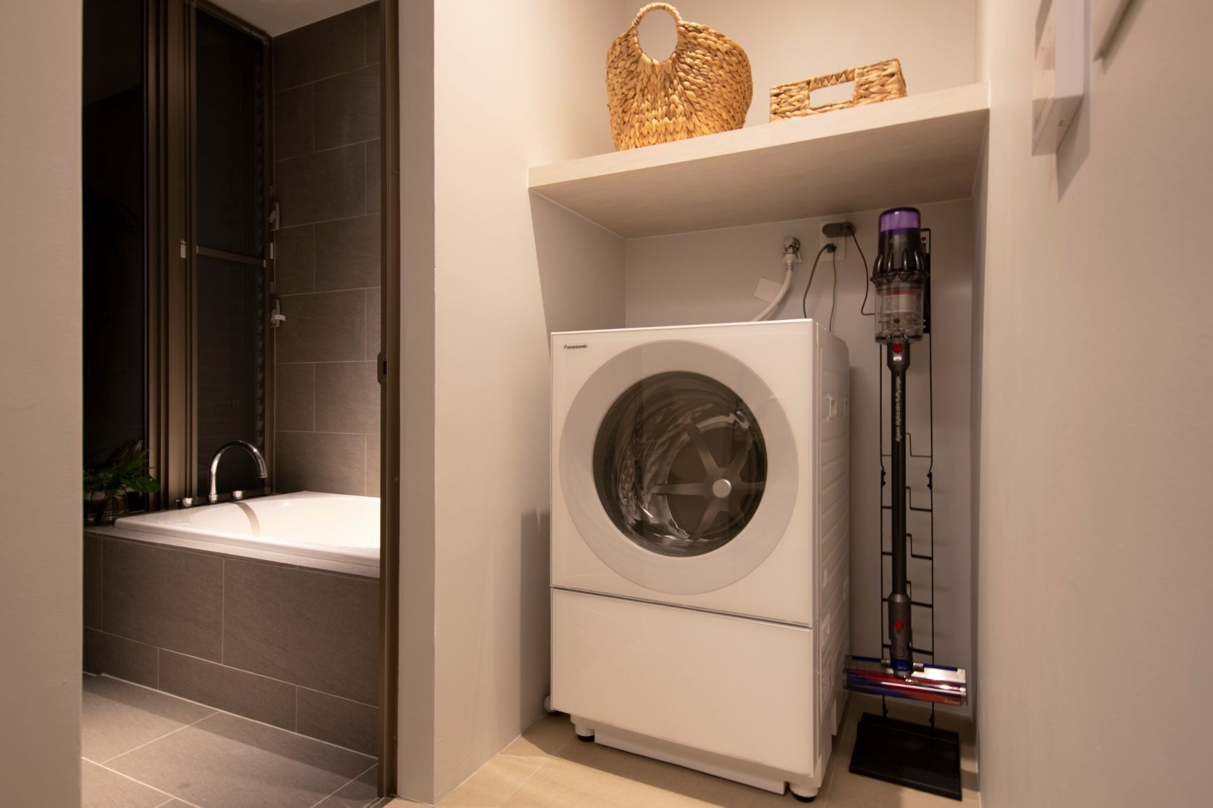 A compact laundry room with a front-loading washing machine, a tall drying machine, and a small shelf with a woven basket and a framed picture.