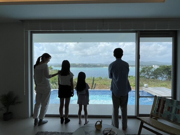 A family standing by a large sliding glass door overlooking a pool and a scenic view of a body of water and hills. - KOURI VILLA Indigo (Nakijin)