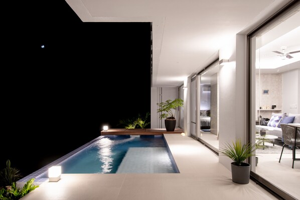 Un área de piscina moderna al aire libre con un ventanal que da a una sala de estar, rodeada de plantas en macetas e interiores bien iluminados.