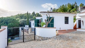 A stone-paved courtyard with white buildings, a gate, and a view of a forested hill. - Spectacular Views Private Villa Valtocado (Mijas Pueblo)