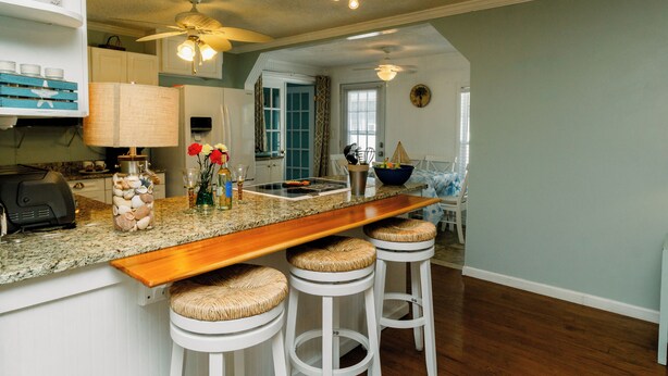 A kitchen with a wooden bar, white bar stools, a ceiling fan, and a view into another room. - PRIVATE Pool w/ Lithium  Golf Cart w paid parking INCLUDED. Walk to Restaurants (Oak Island)