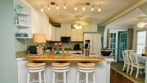 A kitchen with a central island, white bar stools, a ceiling fan, and a dining area with chairs and a table. - PRIVATE Pool w/ Lithium  Golf Cart w paid parking INCLUDED. Walk to Restaurants (Oak Island)